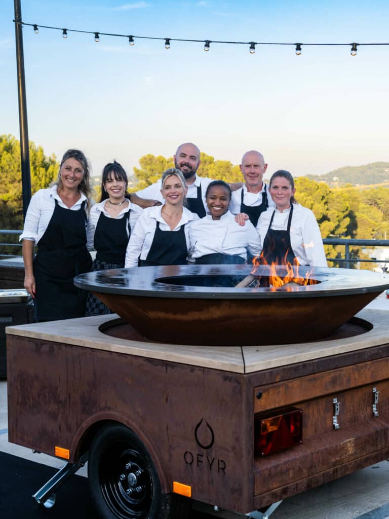 Barbecue OFYR : Équipe de Chefs pour Cuisine Extérieure Équipe de chefs souriants devant un braséro OFYR en extérieur sur une terrasse au coucher du soleil.