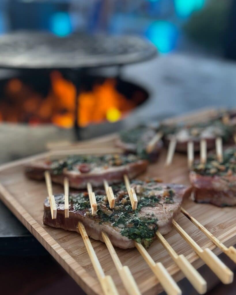 Grillades au Barbecue : Brochettes à la Sauce Chimichurri Brochettes de viande à la sauce chimichurri sur planche en bois, devant un feu de barbecue crépitant.