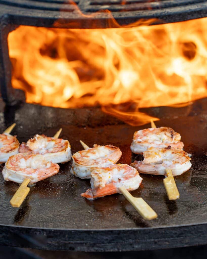 Cuisson de brochettes de crevettes grillées au feu de bois Brochettes de crevettes grillant sur une plaque de cuisson circulaire devant des flammes vives de feu de bois.