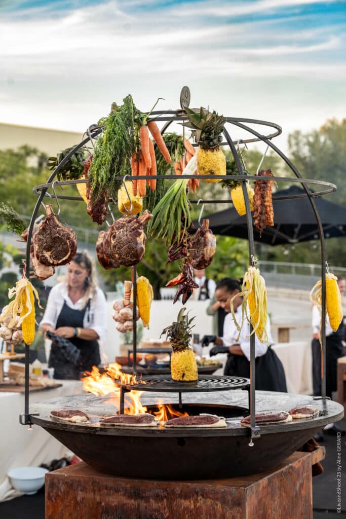 Gastronomie en plein air : Cuisson spectaculaire au brasero Cuisson au feu de bois de viandes et légumes suspendus sur un brasero design lors d'un événement culinaire en plein air.