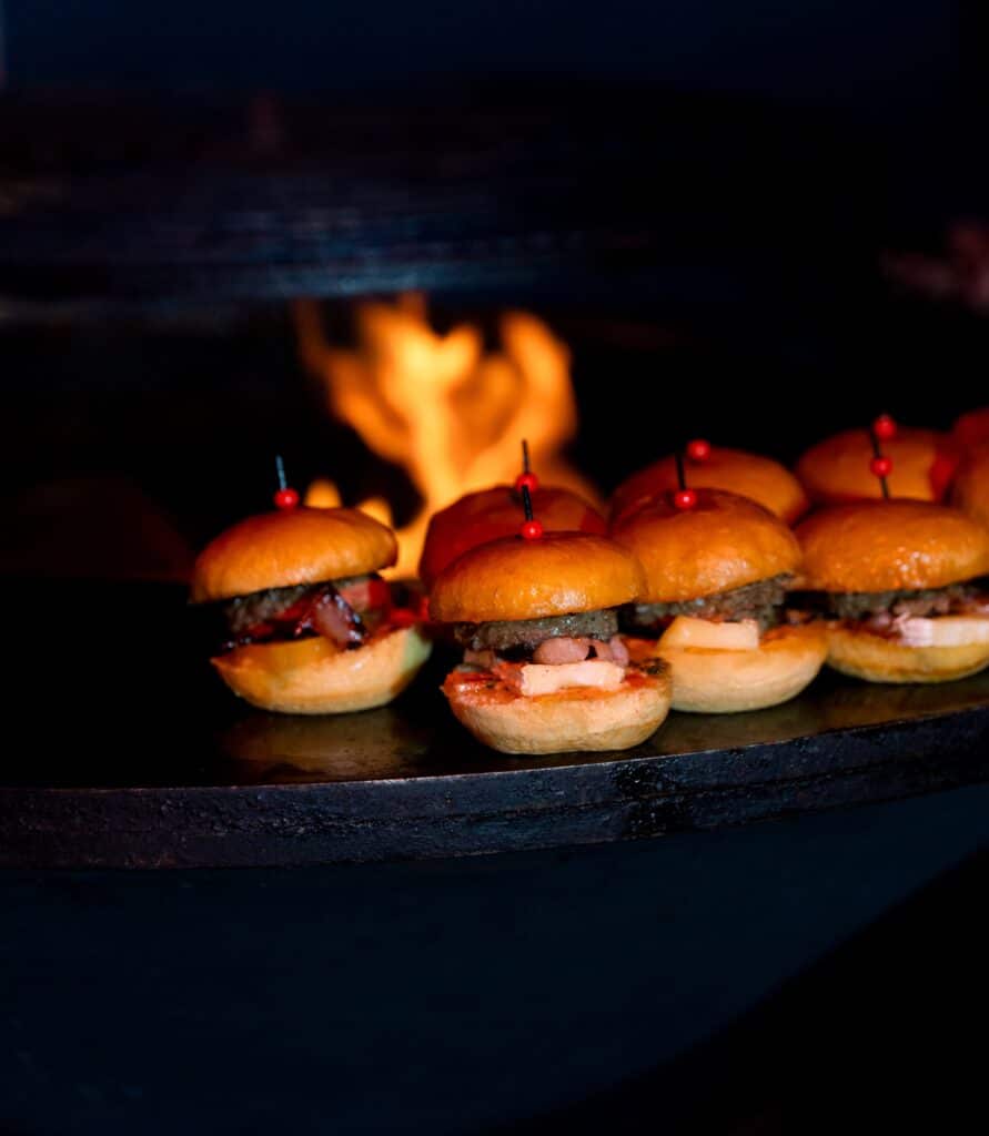 Mini burgers grillés au feu de bois pour vos soirées Mini burgers appétissants sur une plancha devant des flammes de feu de bois, ambiance de cuisine chaleureuse.