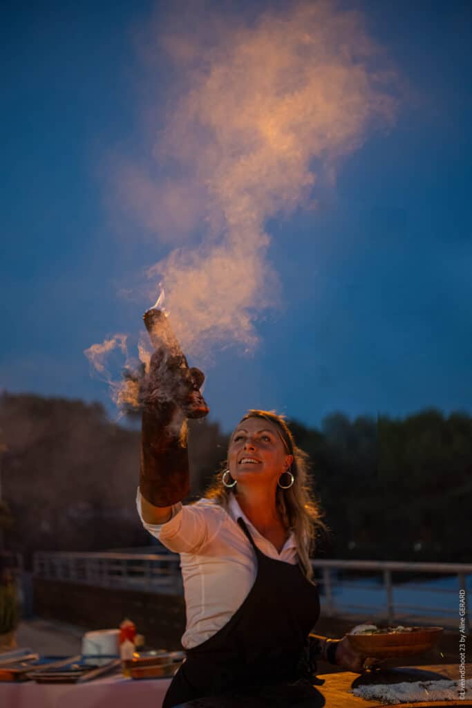 Démonstration de cuisine au flambadou par une femme chef Chef souriante maniant un flambadou ardent lors d'une démonstration culinaire nocturne en plein air.