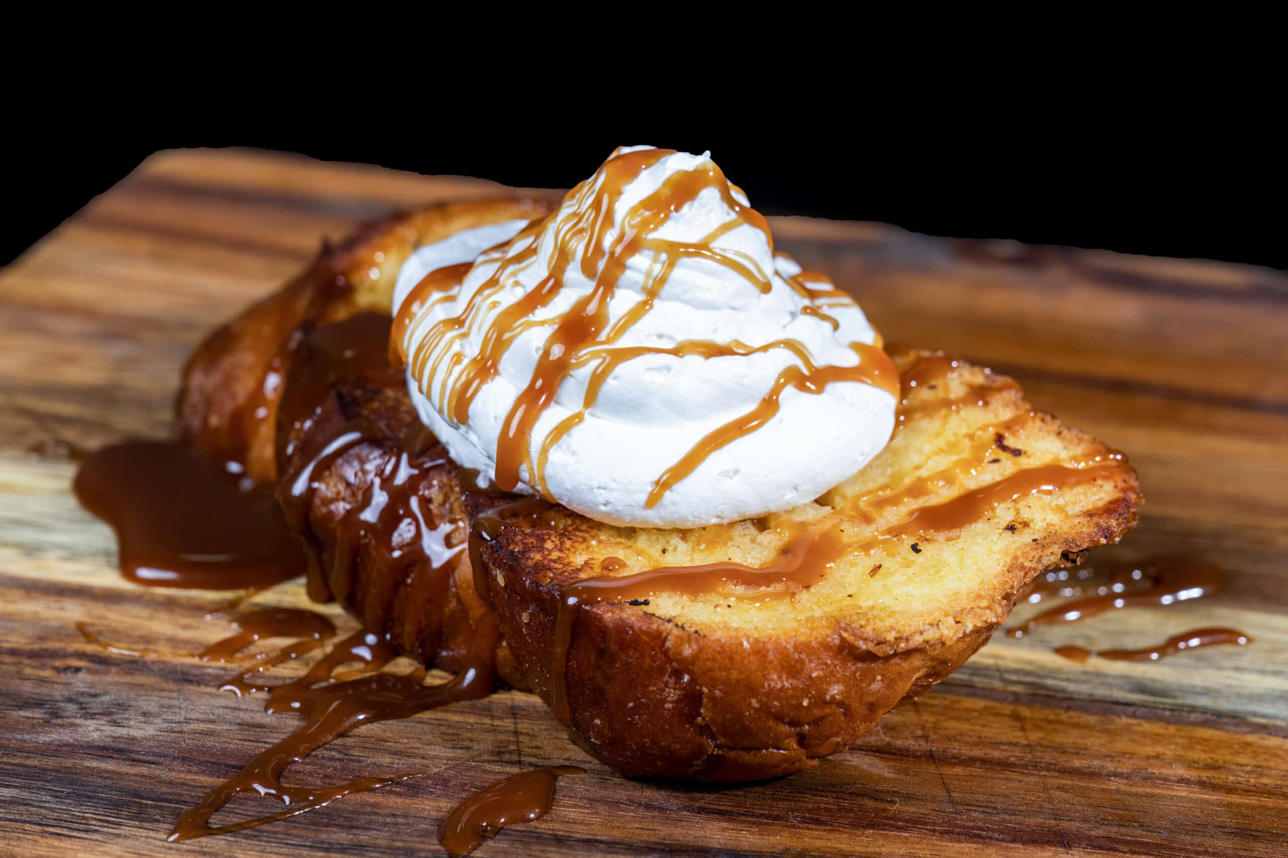 Pain Perdu Brioché, Crème Fouettée et Coulis de Caramel Pain perdu brioché doré avec crème fouettée et coulis de caramel fondant servi sur une planche en bois.